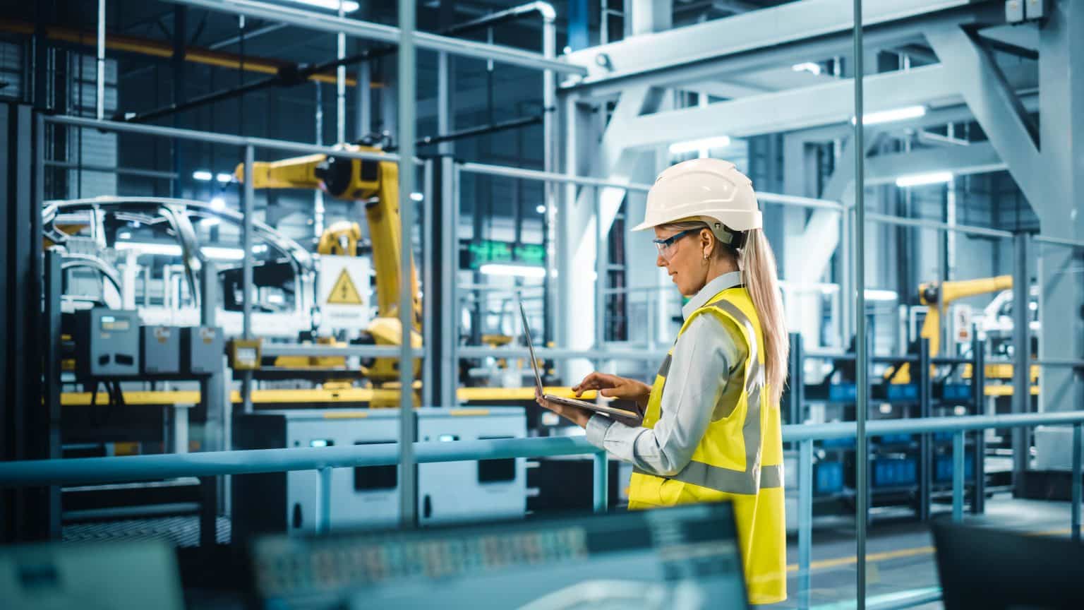 Car Factory: Female Automotive Engineer Wearing Hard Hat, Standing, Using Laptop. Monitoring, Control, Equipment Production. Automated Robot Arm Assembly Line Manufacturing Electric Vehicles.